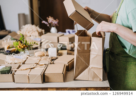Person arranges kraft gift bags and handmade soap boxes at a worktable filled with natural decor. Creative entrepreneur, artisan packaging, brand authenticity, local business 126949914