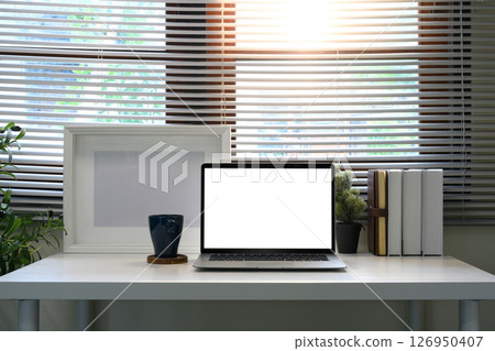 Home office desk with a blank screen laptop, coffee mug, books, and plants near a window. 126950407