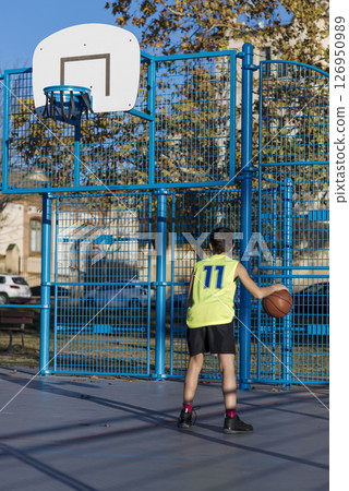 Boy dribbling basketball on outdoor court with blue fence. 126950989