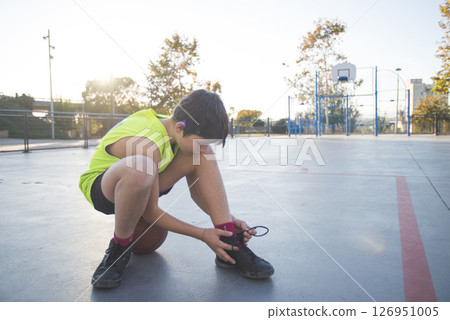 Boy tying his shoe on an outdoor basketball court at sunset. 126951005