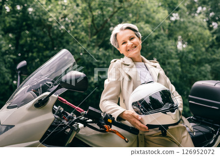Smiling senior woman with gray hair holding helmet near motorcycle, enjoying freedom and confidence 126952072