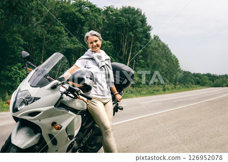 Happy mature woman with gray hair enjoying freedom on motorcycle beside scenic countryside road 126952078