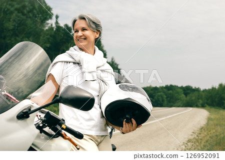 Smiling senior woman with gray hair and helmet standing by motorcycle on open road, summer freedom 126952091