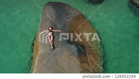 A person lies peacefully on a smooth rock surrounded by crystal-clear water, soaking up the sun. The serene atmosphere captures the essence of a perfect day by the sea. 126952294