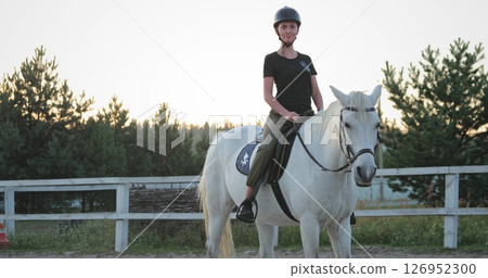 Horsewoman wearing a helmet gracefully riding a stunning white horse in a paddock at sunset, surrounded by a rustic wooden fence and the tranquil beauty of nature 126952300