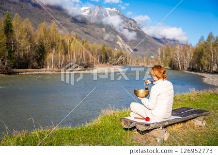 Woman playing a Tibetan singing bowl by a mountain river in Altai. Concept of sound healing, spiritual alignment, and deep connection with natural elements Woman playing a Tibetan singing bowl by a mountain river in Altai. Concept of sound healing, spiritual alignment, and deep connection with natural elements 126952773