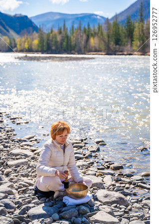 Woman performing a sound ritual with a Tibetan singing bowl by the Altai river. Concept of feminine energy, sacred vibration, and shamanic connection to the Earth 126952777