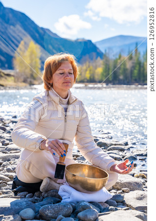 Woman performing a sound ritual with a Tibetan singing bowl by the Altai river. Concept of feminine energy, sacred vibration, and shamanic connection to the Earth 126952816