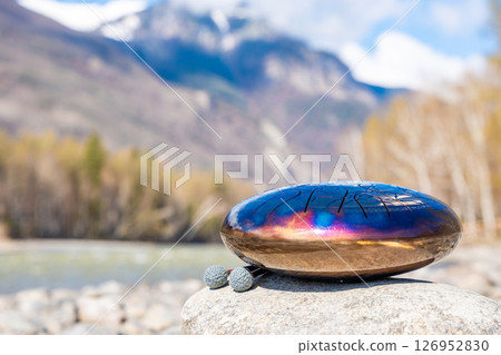 Steel tongue drum resting on a stone by the river in the Altai mountains. Concept of elemental harmony, raw sound energy, and nature as a sacred space for music and silence 126952830