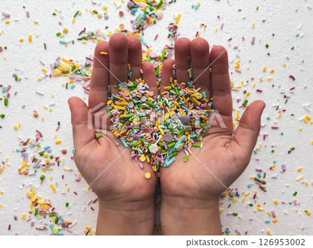 Close-up of child's hands playing with colorful sprinkles on table. Heart-shaped sprinkles in a child's hands. 126953002