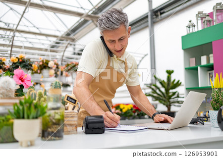 A Caucasian male shop owner takes an order over the phone while working on his computer at his flower shop. A Caucasian male shop owner takes an order over the phone while working on his computer at his flower shop. 126953901