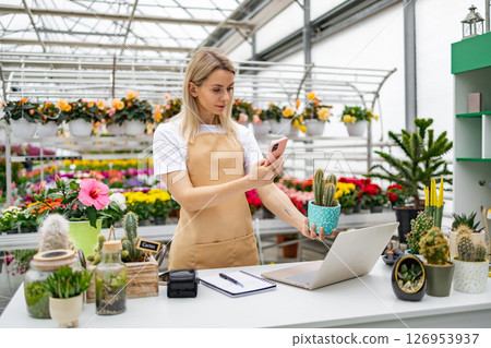 A florist is capturing a photo of a cactus while working in her shop using a smartphone and a laptop. 126953937