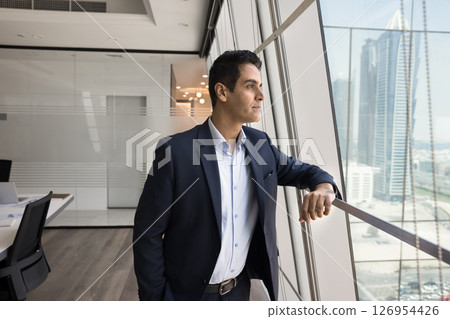 Thoughtful Indian businessman standing near window and looking at city Thoughtful Indian businessman standing near window and looking at city 126954426