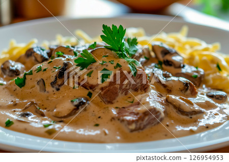 Close-up of classic beef stroganoff with tender beef, creamy mushroom sauce, and fresh parsley, served over pasta on a white plate in a cozy setting 126954953