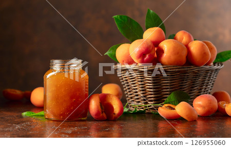 Apricot jam in glass jar and fresh fruits on a brown table. 126955060