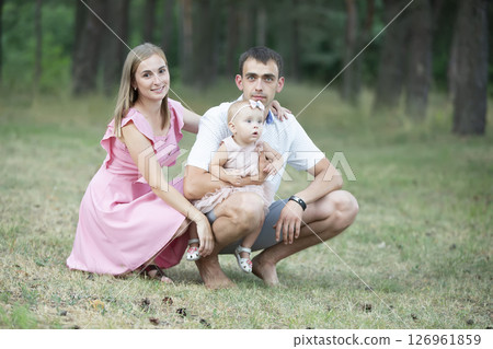 Husband, wife and their little daughter for a walk in the park. Happy dad and mom with their one-year-old daughter are looking at the camera. Husband, wife and their little daughter for a walk in the park. Happy dad and mom with their one-year-old daughter are looking at the camera. 126961859