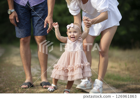 Happy one-year-old girl is being held by dad and mom. Little child walking with parents. A cute baby learns to walk with the help of her parents. 126961863