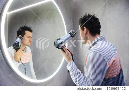 A man with damp hair styles himself using a hairdryer while looking into a round LED-lit mirror in a clean, modern bathroom interior. A man with damp hair styles himself using a hairdryer while looking into a round LED-lit mirror in a clean, modern bathroom interior. 126963422