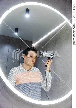 Young man stands in front of a glowing round mirror, carefully drying and styling his wet hair with a hairdryer in a modern bathroom. Young man stands in front of a glowing round mirror, carefully drying and styling his wet hair with a hairdryer in a modern bathroom. 126963426
