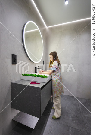Teenage girl in floral pajamas turns off water after washing hands in stylish bathroom with round mirror and green decor Teenage girl in floral pajamas turns off water after washing hands in stylish bathroom with round mirror and green decor 126963427