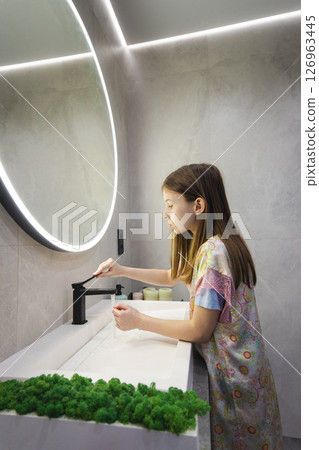 Teen girl in colorful nightgown turns off faucet after washing hands in modern bathroom with large mirror and LED lighting 126963445