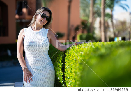 Woman in white dress standing in front of green bush on vacation Woman in white dress standing in front of green bush on vacation 126964389