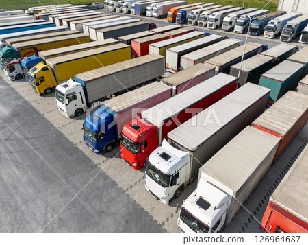 Aerial view of a large truck parking lot at a logistics hub, with multiple commercial semi trucks parked in organized rows. Aerial view of a large truck parking lot at a logistics hub, with multiple commercial semi trucks parked in organized rows. 126964687