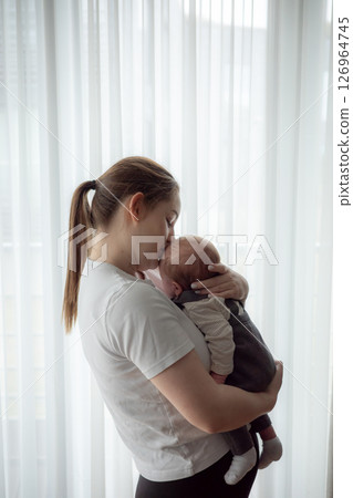 Young mother holding newborn baby in soft natural light standing by a window at home. Concept of early motherhood, tenderness, bonding, parental love, and infant care in a calm home environment. 126964745