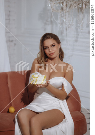 Woman in white dress sitting on pink sofa holding birthday cake with candle in classic room 126968074