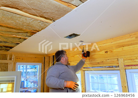 Worker is securing drywall to wooden beams while wearing gloves handling drill inside residential space. Worker is securing drywall to wooden beams while wearing gloves handling drill inside residential space. 126968456