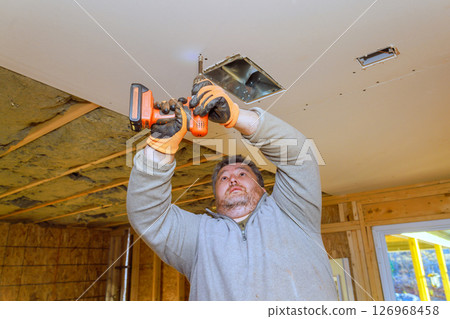 Man uses drill installing drywall to ceiling in renovation project at indoor construction site. 126968458