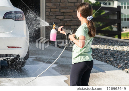 A young girl enthusiastically assists in washing the family's electric car in their suburban driveway. A young girl enthusiastically assists in washing the family's electric car in their suburban driveway. 126968649