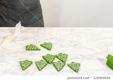 A workspace featuring rows of green Christmas tree cookies, ready for decorating, with decorating tools visible in the background. A workspace featuring rows of green Christmas tree cookies, ready for decorating, with decorating tools visible in the background. 126968688