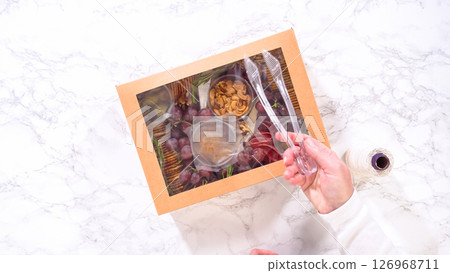 Flat lay. Hands are meticulously adding fresh red grapes to a bowl, complementing a beautifully arranged charcuterie box brimming with a variety of cheeses, olives, and cured meats, set against a Flat lay. Hands are meticulously adding fresh red grapes to a bowl, complementing a beautifully arranged charcuterie box brimming with a variety of cheeses, olives, and cured meats, set against a 126968711