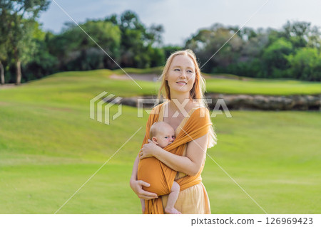 Mother holding her little baby in a yellow sling in the park. Warm and loving family moment. Babywearing, parenting, and mother-child bonding concept Mother holding her little baby in a yellow sling in the park. Warm and loving family moment. Babywearing, parenting, and mother-child bonding concept 126969423
