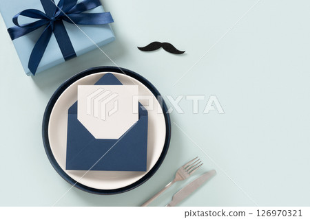 Creative table setting for Father Day. Dining plate with blue envelope and white postcard with gift on blue background. View from above. Flat lay. Copy space 126970321