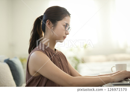 一位二十多歲的女士戴著眼鏡，坐在客廳裡，面對著一台筆記型電腦 126971570