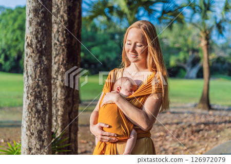 Mother holding her little baby in a yellow sling in the park. Warm and loving family moment. Babywearing, parenting, and mother-child bonding concept 126972079