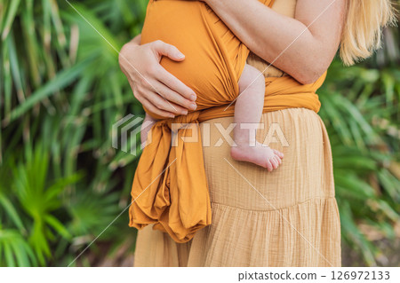 Mother holding her little baby in a yellow sling in the park. Warm and loving family moment. Babywearing, parenting, and mother-child bonding concept Mother holding her little baby in a yellow sling in the park. Warm and loving family moment. Babywearing, parenting, and mother-child bonding concept 126972133