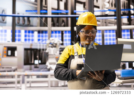 Professional engineer in warehouse, focused on maintenance of solar energy systems and machinery. African american woman with laptop ensuring optimal performance and promoting sustainable development. 126972333