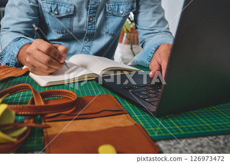 Craftsman working on laptop and taking notes in notebook, running small business Craftsman working on laptop and taking notes in notebook, running small business 126973472
