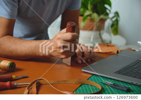 Shoemaker working leather with craft tools and laptop in his workshop 126973600