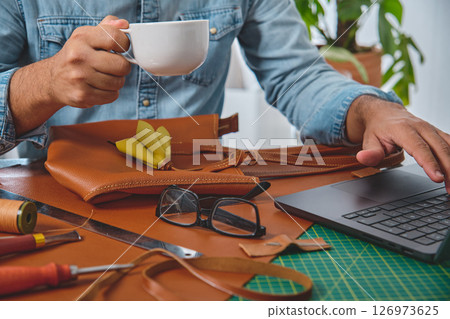 Leather craftsman drinking coffee and working on laptop in his workshop 126973625