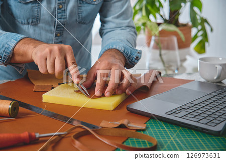 Craftsman cutting leather in his workshop with laptop and tools Craftsman cutting leather in his workshop with laptop and tools 126973631