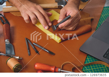 Shoemaker working leather with tools and laptop in workshop 126973635