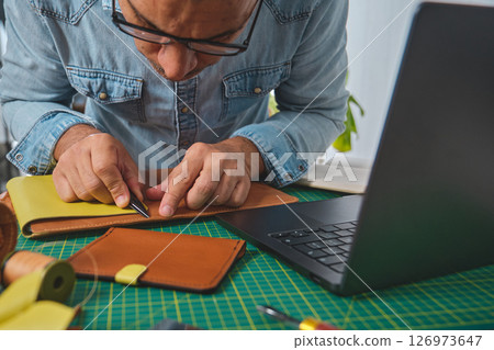 Craftsman working leather at desk with laptop and tools Craftsman working leather at desk with laptop and tools 126973647