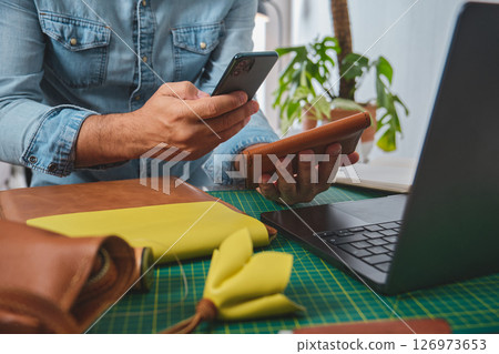 Leather craftsman managing online orders with smartphone and wallet in his workshop 126973653