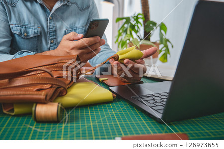 Leather craftsman working with leather goods at a workshop using smartphone and laptop Leather craftsman working with leather goods at a workshop using smartphone and laptop 126973655