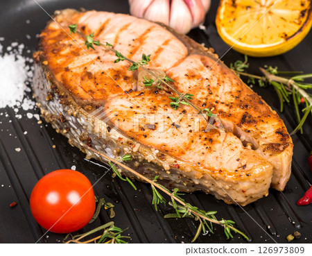 AI generated top view of succulent sea bass steak pieces grilled over charcoal, perfectly cooked with light smoke. Seasoned with salt and black pepper, garnished with fresh rosemary and lemon slices. 126973809