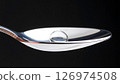 A macro shot of a single drop of liquid or a clear pill on a silver spoon against a black background. 126974508